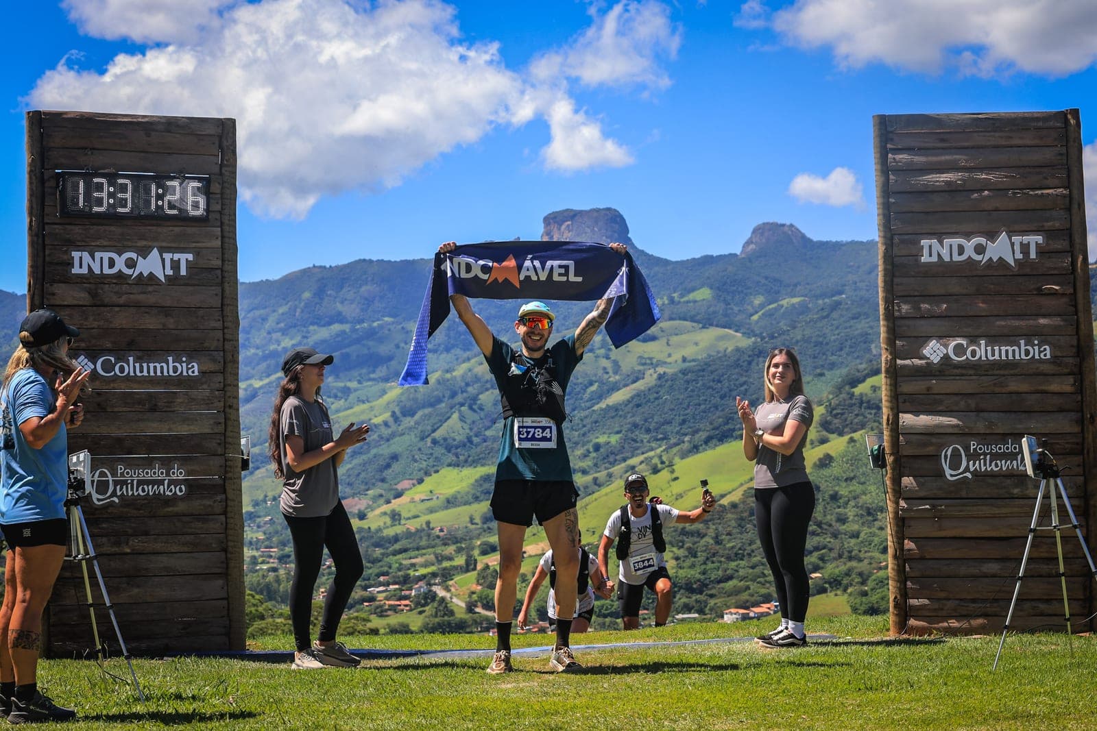 Felipe Bessa comemorando de braços erguidos na chegada de uma ultra de trail, entre montanhas.